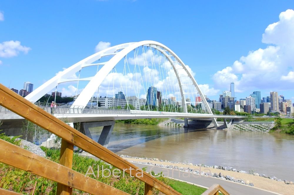 A white arch bridge spans a river with a city skyline, including tall buildings in the background, under a clear blue sky. Stairs and greenery are in the foreground. Text reads "Best Places to Live in Alberta, Canada.