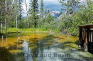 A natural pool with clear water surrounded by trees and mountains, with a wooden structure partially visible on the right. "Cave and Basin National Historic Site" text is at the bottom.