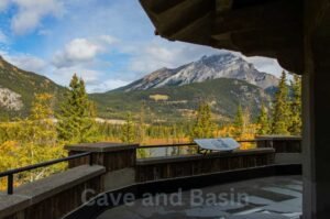 View from a wooden observation deck overlooking a mountainous landscape with lush greenery and a signboard. The foreground is shaded while the background is brightly lit. The words "Cave and Basin National Historic Site" are visible.