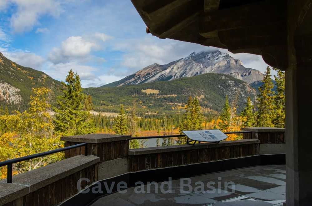 View from a wooden observation deck overlooking a mountainous landscape with lush greenery and a signboard. The foreground is shaded while the background is brightly lit. The words "Cave and Basin National Historic Site" are visible.