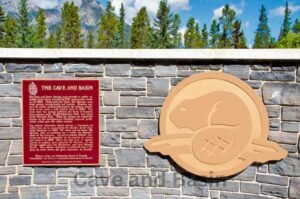 A sign and a beaver emblem are proudly mounted on a stone wall at the Cave and Basin National Historic Site, with majestic mountains and lush trees creating a breathtaking backdrop.