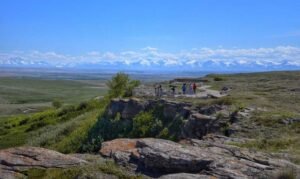 Tourists stand on a viewing platform overlooking the vast, green landscape of Head-Smashed-In Buffalo Jump with distant snow-capped mountains under a clear blue sky.