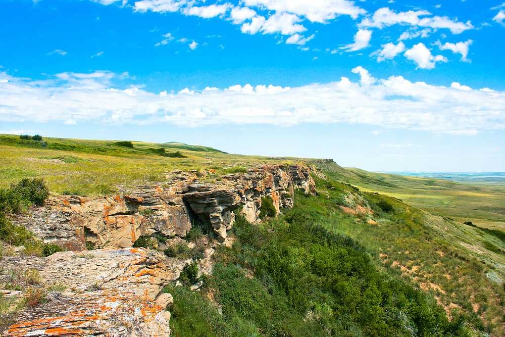 A grassy plateau with rocky cliffs and a vast, open plain under a blue sky with scattered clouds, reminiscent of the historic Head-Smashed-In Buffalo Jump.