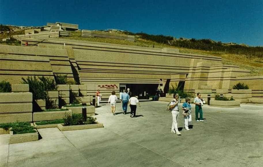 A group of people stands outside a large, modernist concrete building with tiered structures and landscaping, reminiscent of the ancient Head-Smashed-In Buffalo Jump, enjoying a sunny day.