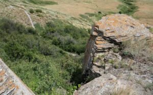 Large rock formations with visible layers overlook a green, shrubbery-filled valley at Head-Smashed-In Buffalo Jump. A dirt trail winds through the ancient landscape.