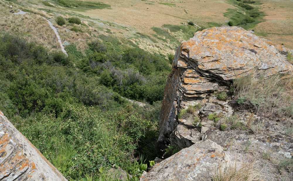 Large rock formations with visible layers overlook a green, shrubbery-filled valley at Head-Smashed-In Buffalo Jump. A dirt trail winds through the ancient landscape.