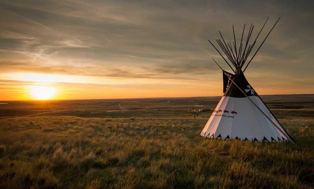 A teepee stands on a grassy field during sunset, with the sun low on the horizon and the vast landscape of Head-Smashed-In Buffalo Jump visible in the background.