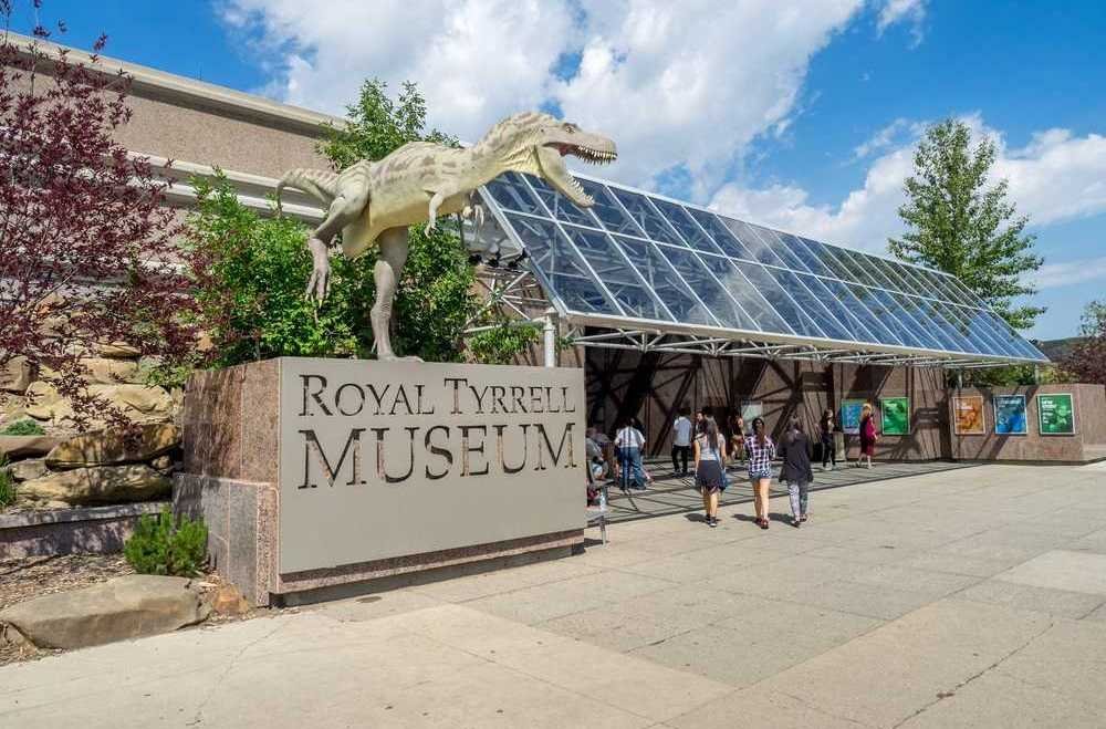 Entrance of the Royal Tyrrell Museum, featuring a dinosaur statue and a group of people walking towards the glass-covered walkway on a sunny day.