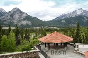 Scenic view of a pavilion with a red-tiled roof in a mountainous landscape surrounded by lush pine trees, near the Cave and Basin National Historic Site, with snow-capped peaks in the background and a cloudy sky overhead.