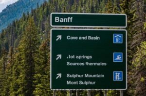 A directional road sign in Banff points toward Cave and Basin National Historic Site, Hot Springs, and Sulphur Mountain, with icons indicating a historical site, swimming, and scenic viewpoint respectively. Forested mountains provide a picturesque background.