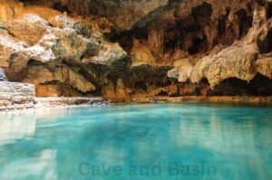 Natural rock cave with stalactites and a clear, turquoise pool of water. The words "Cave and Basin National Historic Site" are visible at the bottom.