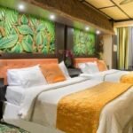 A hotel room with two double beds, vibrant leaf-patterned wall art, and tiki-themed decor. A large window on the right provides a view of a cityscape. Perfect for those seeking hotels near West Edmonton Mall.