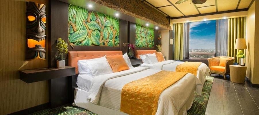 A hotel room with two double beds, vibrant leaf-patterned wall art, and tiki-themed decor. A large window on the right provides a view of a cityscape. Perfect for those seeking hotels near West Edmonton Mall.