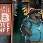 Sign reading "Welcome/Bienvenue Above Banff" next to a cutout of a cartoon bear in a hat and jacket holding a QR code, inviting you to explore the thrilling gondolas in Banff.