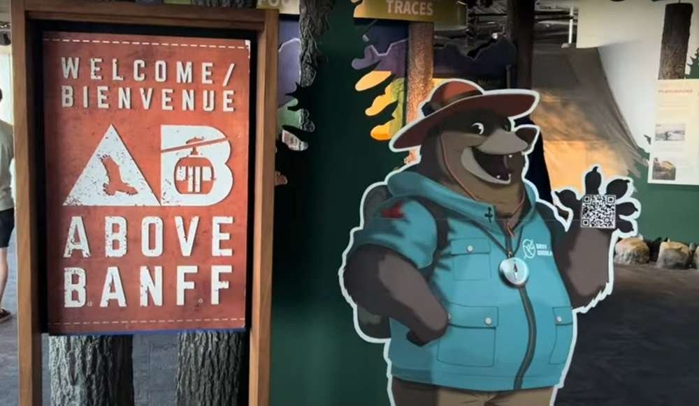 Sign reading "Welcome/Bienvenue Above Banff" next to a cutout of a cartoon bear in a hat and jacket holding a QR code, inviting you to explore the thrilling gondolas in Banff.