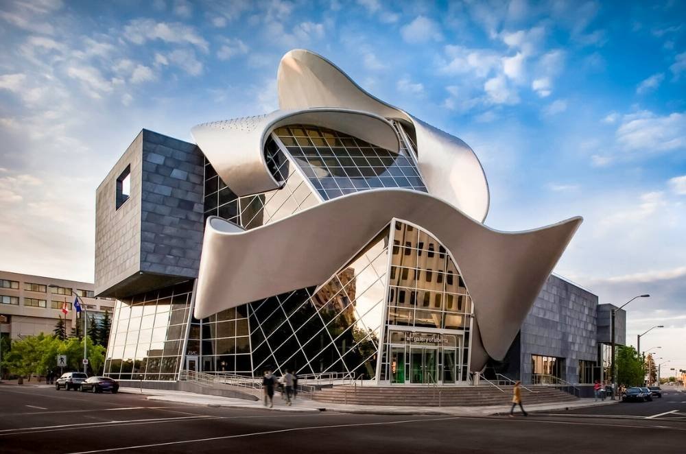 A modern building with a unique, abstract design featuring curved and angular elements. The exterior includes large glass panels, metal structures, and a mix of textures. People are walking by, taking in one of the many free things to do in Edmonton.