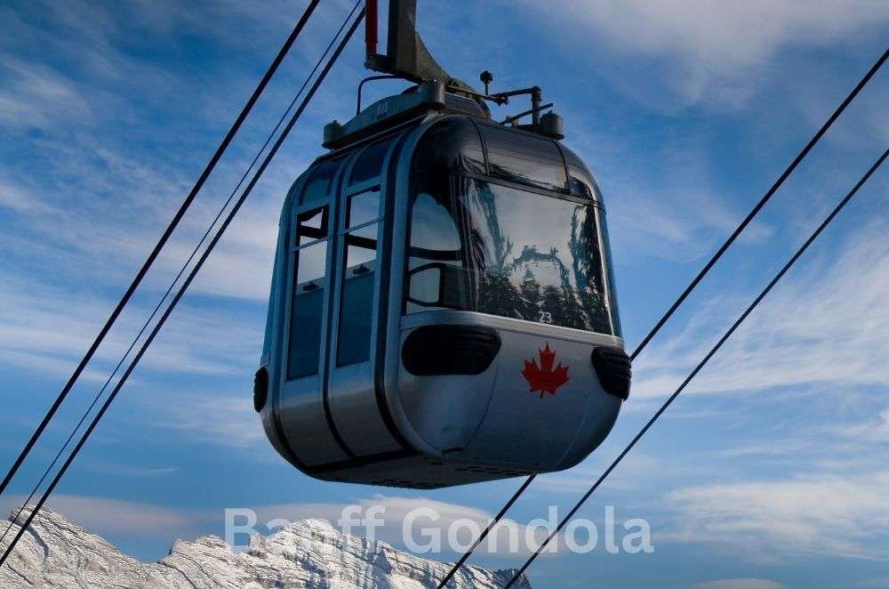 A gondola cabin with a red maple leaf on its side travels along cables against a backdrop of snow-covered mountains and a blue sky. The text "Banff Gondola" is visible at the bottom of the image, showcasing one of the iconic gondolas in Banff.