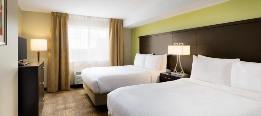 Hotel room with two double beds, a green accent wall, a TV on a dresser, a nightstand with a lamp, and a window with gold curtains. The room is brightly lit with natural and artificial light, perfect for guests seeking hotels near West Edmonton Mall.