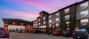 A four-story hotel with a covered entrance and parked cars in the lot at sunset, conveniently located among the finest hotels near West Edmonton Mall.