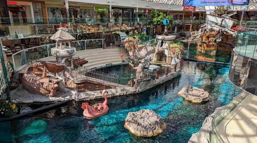A vibrant West Edmonton Mall features an indoor canal with a pirate ship, decorative rocks, and water sculptures, including an island-themed area and a red swan pedal boat floating in blue water.