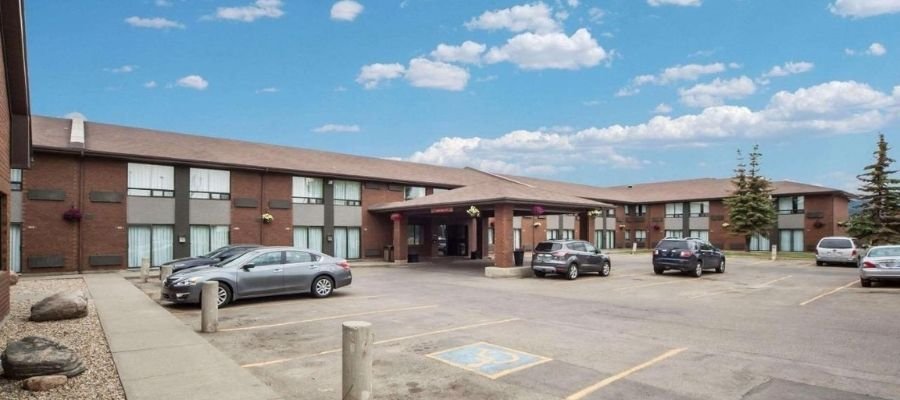 A two-story motel with a brown roof displays several guest rooms and entrances, surrounded by a parking lot with parked cars. The sky is partly cloudy. This cozy spot is among the hotels near West Edmonton Mall, making it convenient for shopping and entertainment.