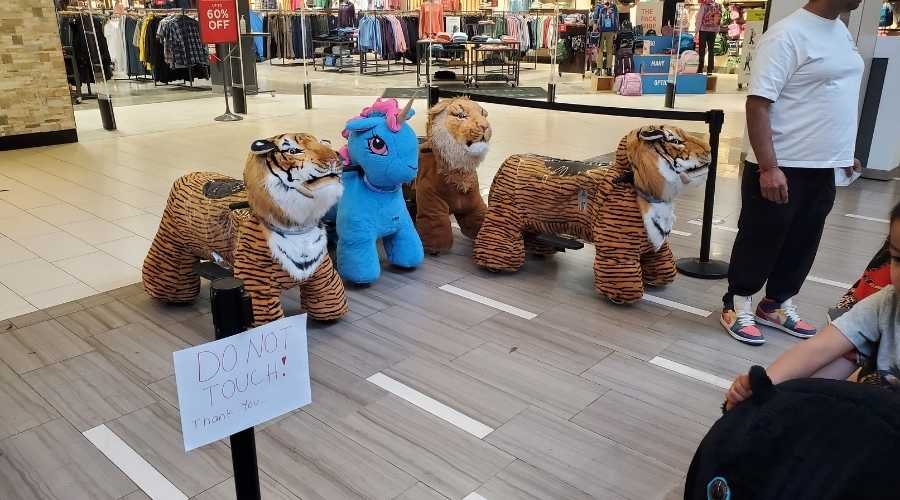 Stuffed tiger and blue unicorn ride-on toys are lined up in West Edmonton Mall, guarded by a "Do Not Touch" sign.