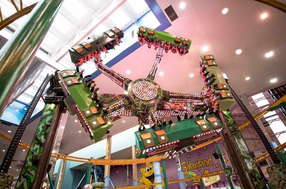 A thrilling amusement park ride at Galaxyland in West Edmonton Mall, with multiple arms, spins passengers in the air indoors, surrounded by bright lights and colorful decor.