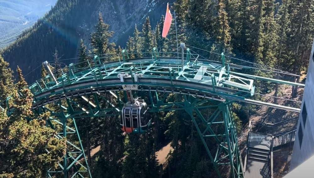 A gondola in Banff travels along a curving, elevated track through a densely forested mountainous area, with a red flag visible on the structure.