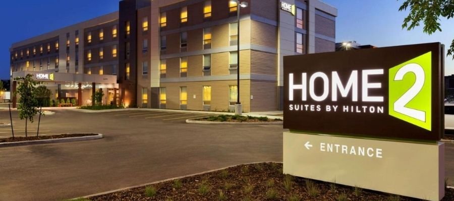 Exterior view of Home2 Suites by Hilton hotel at dusk, featuring a large illuminated sign and landscaped surroundings with parking area and building entrance visible. Conveniently located among hotels near West Edmonton Mall, it's the perfect choice for travelers seeking comfort and accessibility.