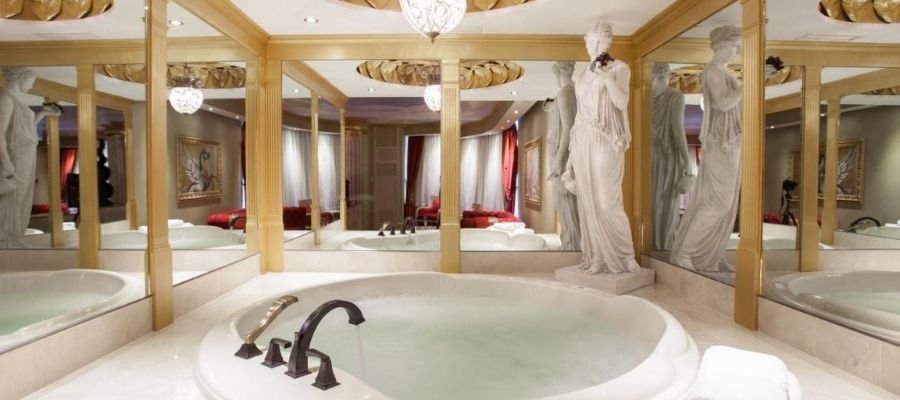 Luxurious bathroom with a large circular bathtub, surrounded by mirrors and adorned with a marble statue. Gold accents and chandeliers add an opulent touch, similar to what you might find in the finest hotels near West Edmonton Mall.