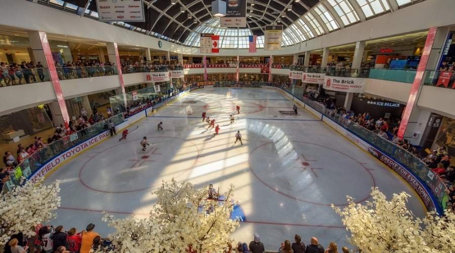 An indoor ice hockey game is being played in West Edmonton Mall. Spectators are watching from multiple levels, while ceiling windows allow natural light to stream in.