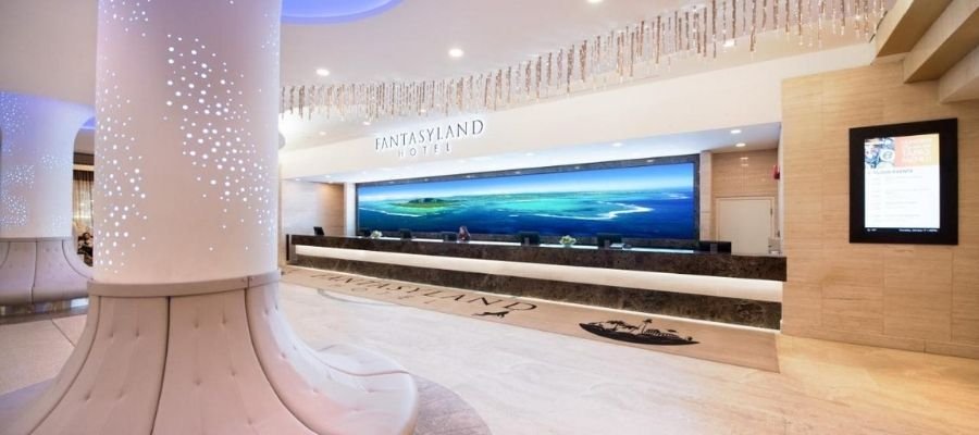 A modern hotel reception area with a long front desk, several receptionists, a large digital screen displaying an ocean view, and a white architectural column in the foreground, reminiscent of hotels near West Edmonton Mall.