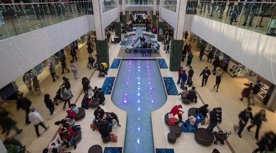 A busy shopping mall interior at West Edmonton Mall features a central water feature and two levels of shops, filled with people walking and sitting on benches.