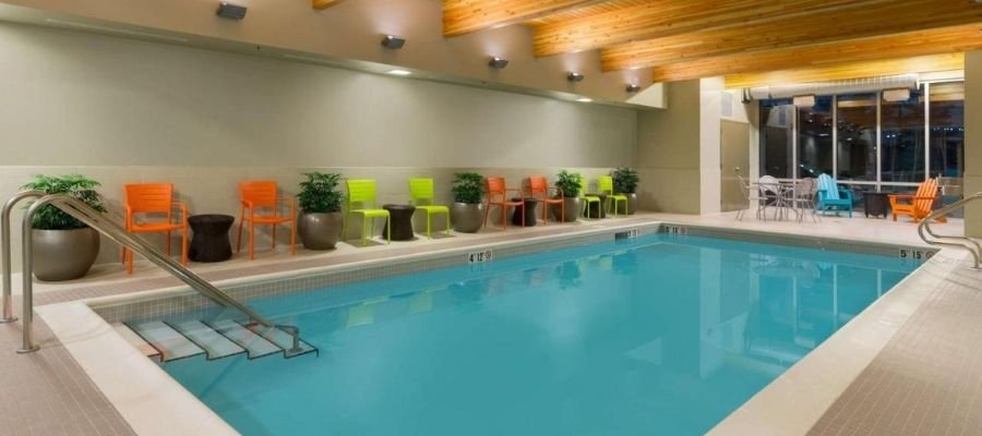 Indoor swimming pool with clear blue water, surrounded by various colored chairs and potted plants. The ceiling has wooden beams, and there are large windows on one side, similar to the serene amenities found at hotels near West Edmonton Mall.