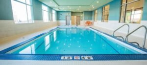 Indoor swimming pool with blue tiles, steps, and depth markers of 1.3 meters and 4 feet 6 inches. Bright windows line the wall, and safety equipment is visible—conveniently located in hotels near West Edmonton Mall.