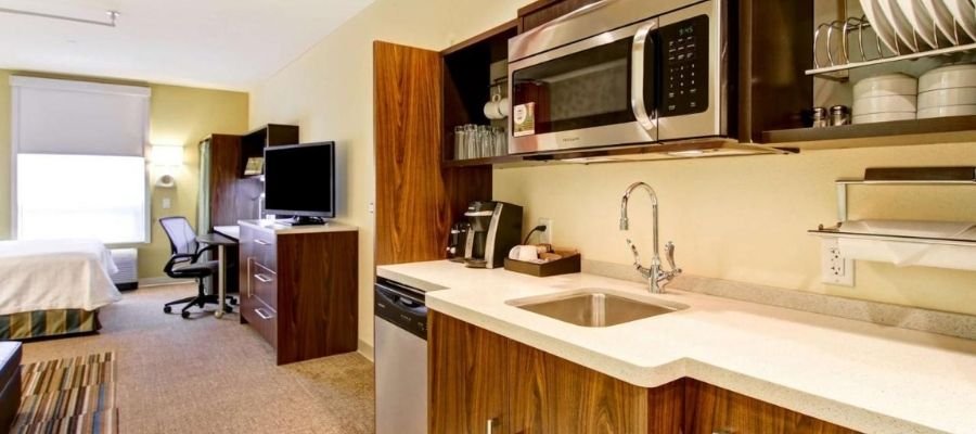 A modern hotel room with a kitchenette, featuring a microwave, sink, and cabinets. The room also includes a TV, desk, and bed. Perfect for your stay in one of the top hotels near West Edmonton Mall.