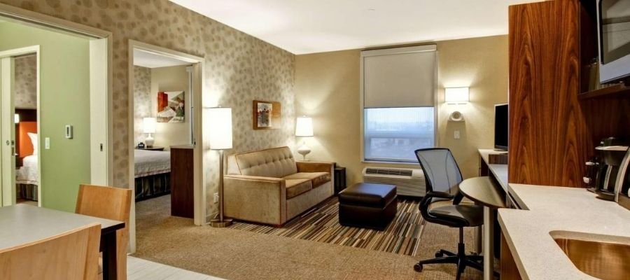 A modern hotel suite near West Edmonton Mall with a living area featuring a sofa, ottoman, desk, chair, and kitchenette. A door leads to a bedroom. The room is well-lit with lamps and natural light from a window.
