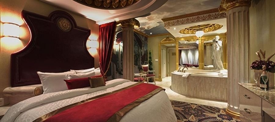 Luxurious hotel room with a large bed, red and gold accents, ornate decor, columns, a round bathtub near a vanity, and detailed ceiling artwork. Perfect for those seeking hotels near West Edmonton Mall.