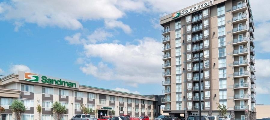 Two Sandman Hotels buildings are shown under a blue sky with scattered clouds. One low-rise building and a taller multi-story structure stand prominently, situated as hotels near West Edmonton Mall. Several cars are parked in front, adding to the bustling atmosphere.