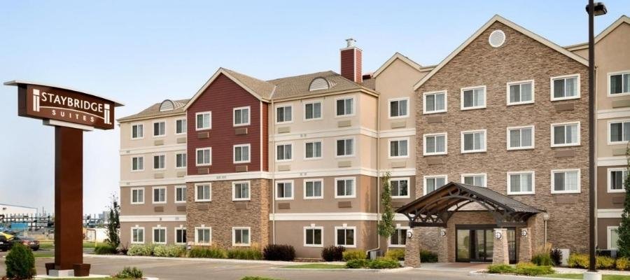 A four-story Staybridge Suites hotel building with a mix of stone and siding exterior, featuring a large sign and covered entrance. Well-maintained landscaping surrounds the entrance area, making it one of the ideal hotels near West Edmonton Mall.