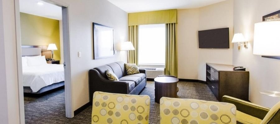 A modern hotel room with a living area featuring a sofa, chairs, coffee table, and a wall-mounted TV. The bedroom area is semi-visible through an open doorway, containing a bed and bedside lamps. Perfect for those seeking hotels near West Edmonton Mall.