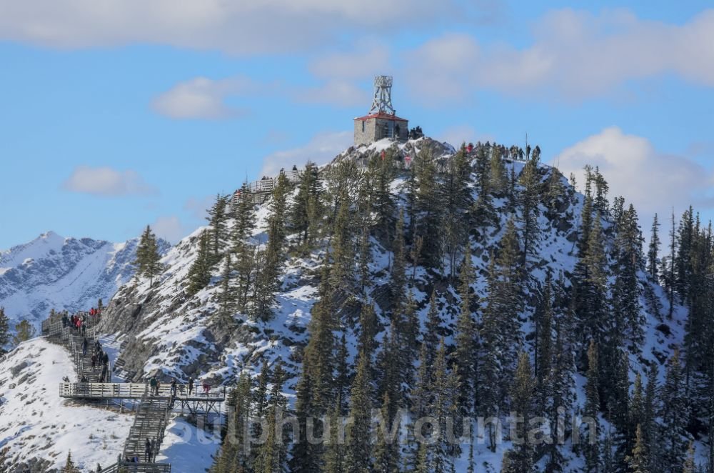 A snowy mountain peak with a small building at the top, surrounded by evergreen trees. A staircase winds up the mountain, and visitors are visible walking along the path, exploring the renowned Sulphur Mountain Trail in Banff National Park.