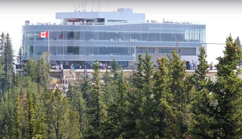 A modern multi-story building with large windows, displaying a Canadian flag, surrounded by trees. Visitors are seen at the lower observation area, reminiscent of the scenic views experienced on gondolas in Banff.
