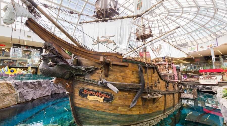 A large pirate ship model with decorative sails is displayed indoors, surrounded by a clear water feature in West Edmonton Mall.