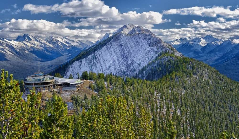 Mountain landscape with a sharp, snow-dusted peak, dense evergreen forest, and a building on a ridge under a partly cloudy sky. Gondolas in Banff glide smoothly above the treetops.