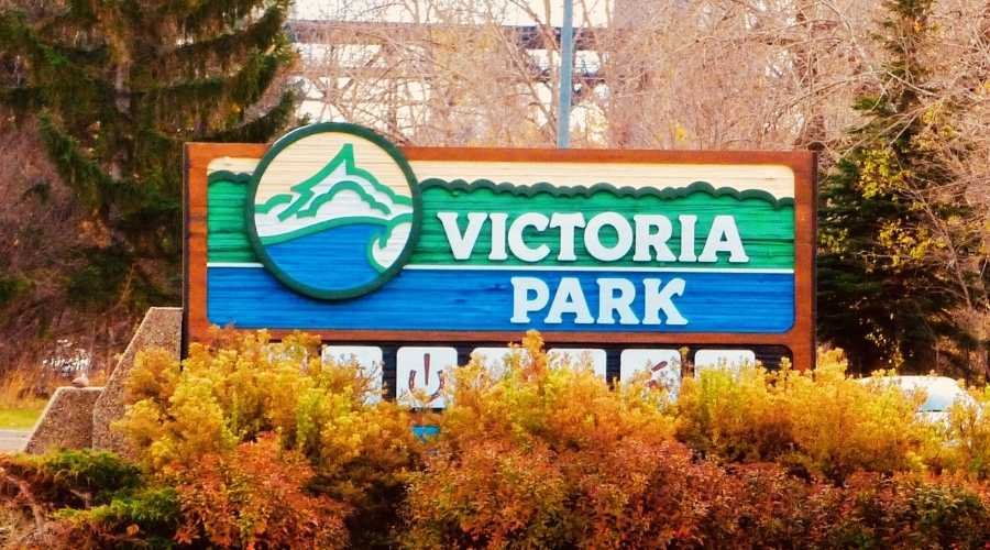 A wooden sign with the text "Victoria Park," one of the best parks in Edmonton, is surrounded by greenery and trees. A bridge is visible in the background.