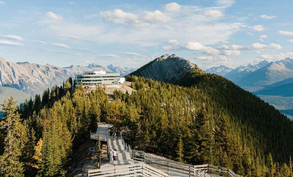 A mountaintop building is surrounded by dense trees with a wooden pathway leading up to it, reminiscent of the scenic routes taken by gondolas in Banff. Rugged mountains are seen in the background under a partly cloudy sky.