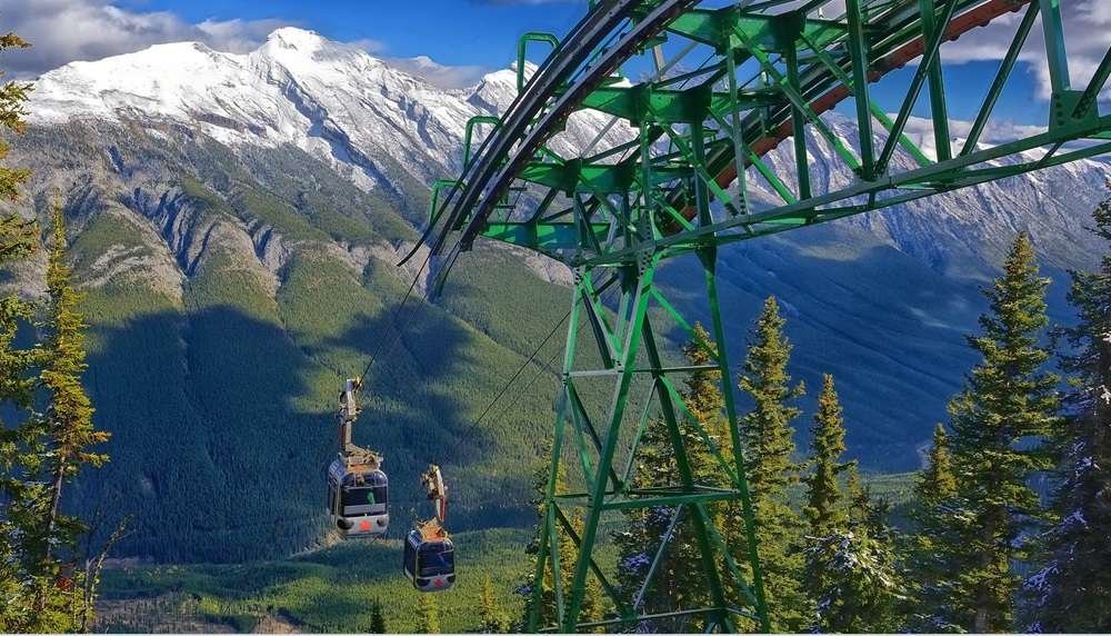 Two gondolas in Banff travel on a mountainous cableway against a backdrop of snow-covered peaks and lush green trees.