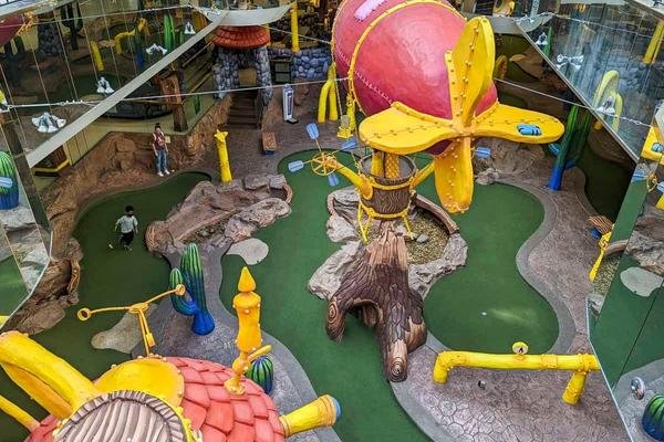 A colorful, whimsical indoor miniature golf course with oversized, cartoonish decorations, including a large red balloon and yellow propellers. Visitors are scattered throughout the course at this delightful West Edmonton Mall attraction.