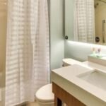 A modern bathroom with a bathtub, white shower curtain, toilet, sink with a large mirror above, a towel rack with white towels, and a framed picture on the wall—reminiscent of the luxurious amenities you might find in hotels near West Edmonton Mall.